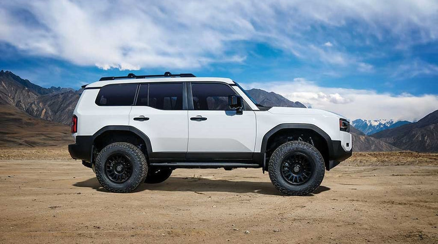 A white Toyota Land Cruiser 250 with lift and black off-road wheels in side profile on a desert plain before mountains.