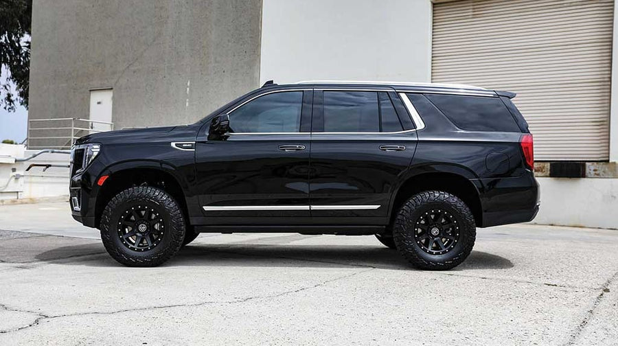 A black leveled GMC Yukon with black ICON wheels and all-terrain tires in side profile beside a warehouse.