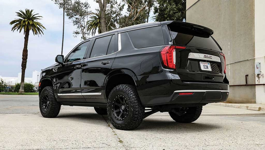 Rear three-quarter of a black leveled GMC Yukon with black ICON wheels and all-terrain tires beside a warehouse and palm trees.