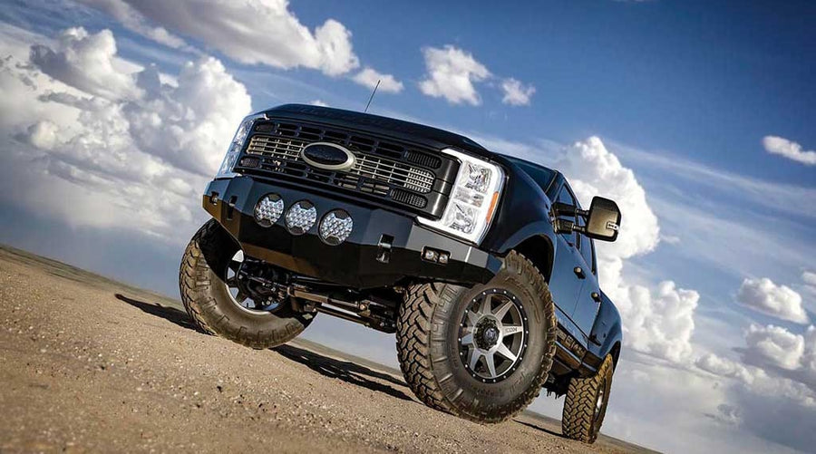 Low front three-quarter of a black lifted Ford Super Duty with steel front bumper and three round lights on a desert flat.
