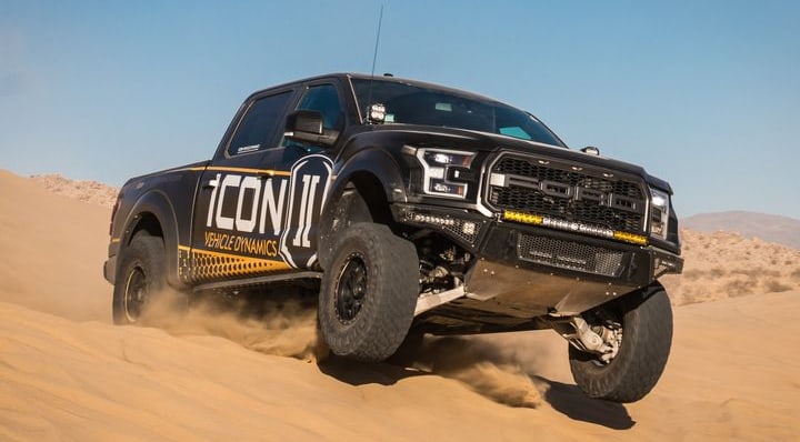 A black second-generation Ford F-150 Raptor with ICON Vehicle Dynamics wrap and light bars kicking up sand in the dunes.