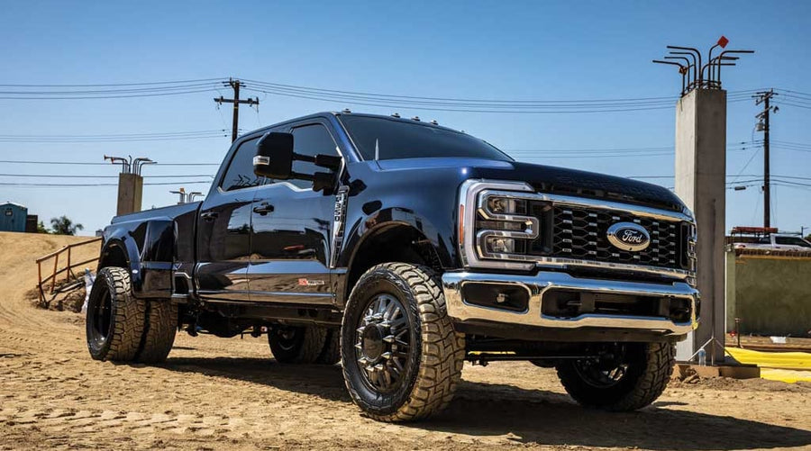 Front three-quarter of a dark blue lifted Ford Super Duty dually crew cab with black wheels and off-road tires on a dirt construction site.