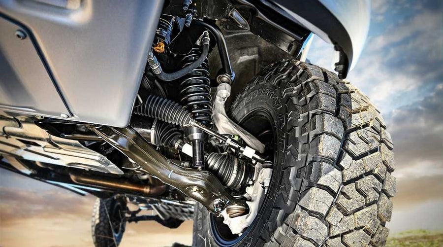 Low close-up of an installed front coilover and control arms behind a large all-terrain tire.