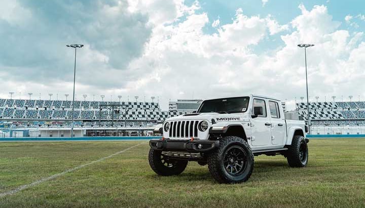 Front three-quarter of a lifted white Jeep Gladiator Mojave on grass in front of stadium grandstands.