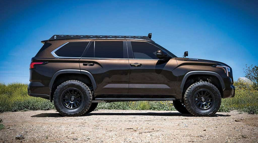 Side profile of a lifted brown Toyota Sequoia with roof rack parked on dirt under a blue sky.