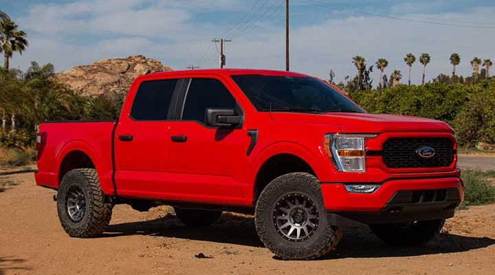 Red lifted Ford F-150 SuperCrew parked on dirt, front three-quarter view, with palms and a rocky hill behind.