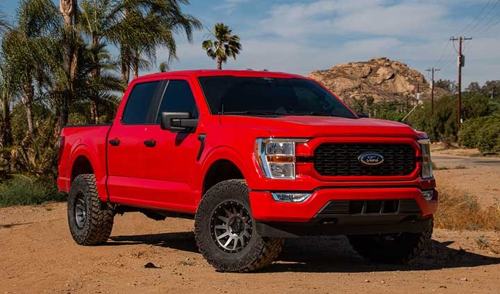 Red lifted Ford F-150 SuperCrew on dirt, front three-quarter from the right.