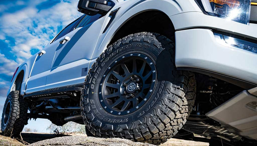 Low-angle view of the front wheel and fender of a white lifted Ford F-150 on a rock under a cloudy sky.