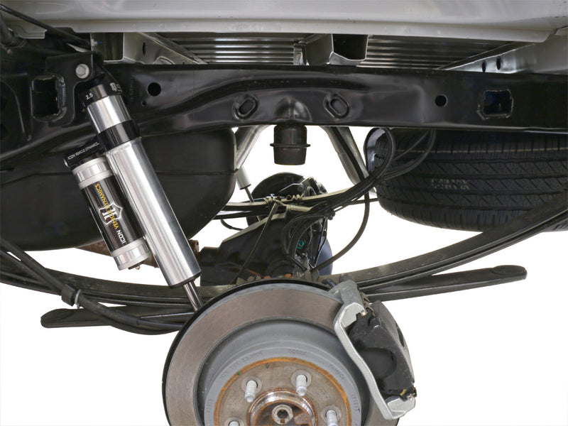 Underbody view of an installed silver ICON 2.5 VS piggyback rear shock beside a rear brake rotor and leaf spring, background cut to white.