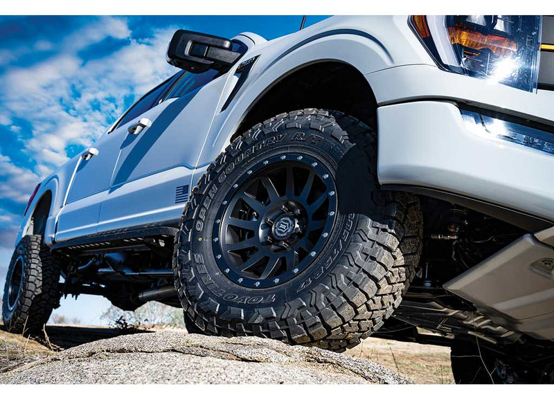 ICON Vehicle Dynamics Stage Suspension System on a Ford F-150: low-angle view of the front wheel and fender of a white lifted style Ford F-150 on a rock under a cloudy sky