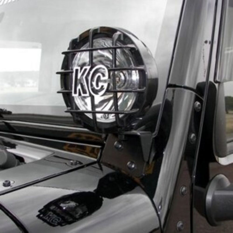 KC HiLiTES A-Pillar Cowl Light Mount Brackets on a Jeep Wrangler JK: round KC light with a stone guard mounted on a black windshield pillar bracket of a black Jeep Wrangler