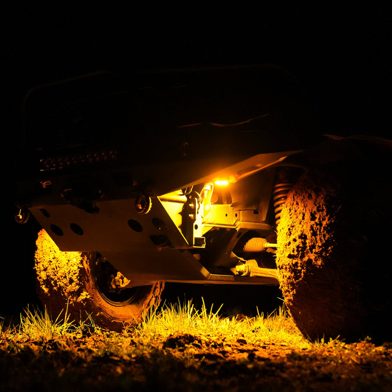 KC HiLiTES Cyclone V2 LED Rock Light 6-Light System on a vehicle: amber rock light glowing under the front of an off-road vehicle at night over muddy tires and grass