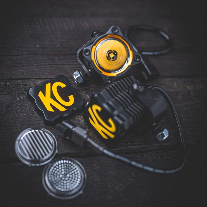 KC HiLiTES FLEX ERA 1 Light Cover & Shield on a vehicle: KC FLEX ERA 1 lights, a black and yellow KC cover and two optic lenses laid out on dark wood
