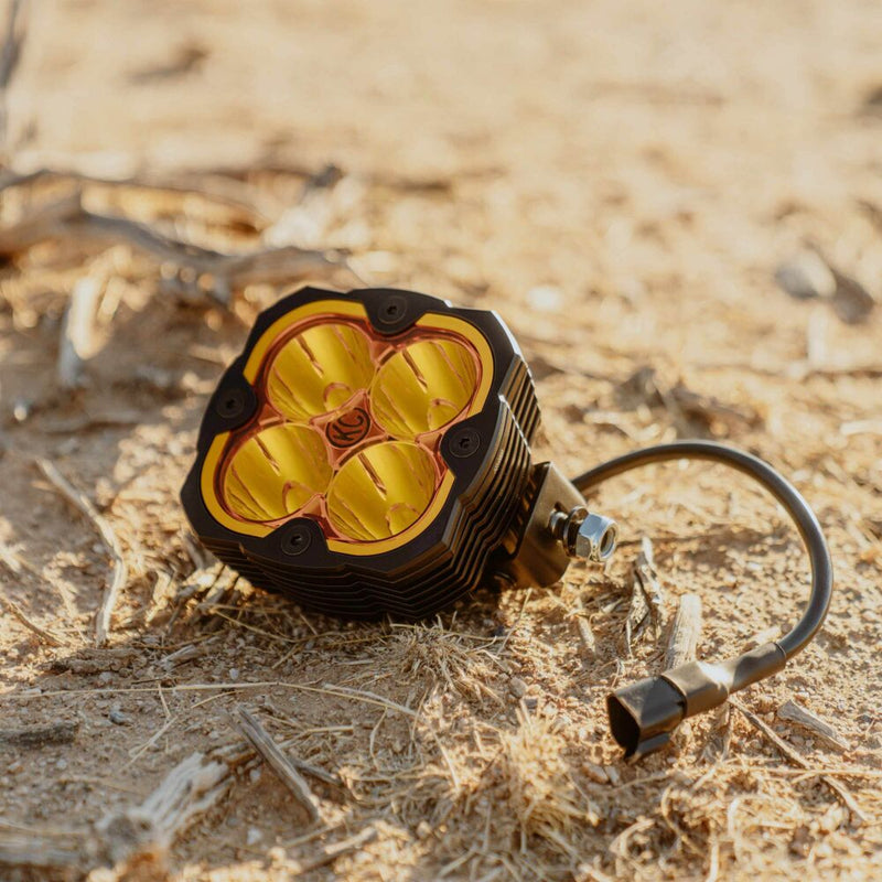 KC HiLiTES FLEX ERA 4 Performance Yellow Lens on a vehicle: FLEX ERA 4 LED light with a yellow four-pod lens resting on dry desert ground with its wiring pigtail