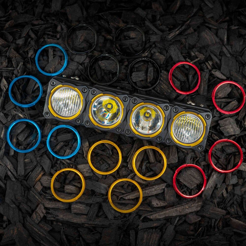 KC HiLiTES FLEX ERA LED Light Bar 10" Segment Bezel Kit on a vehicle: FLEX ERA 10" light bar segment with gold bezel rings, surrounded by loose blue, black, red and gold bezel rings on dark wood mulch