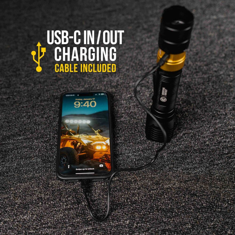 KC HiLiTES FLX 3K Rechargeable Flashlight on a vehicle: flashlight standing on carpet with a USB-C cable charging a smartphone lying next to it