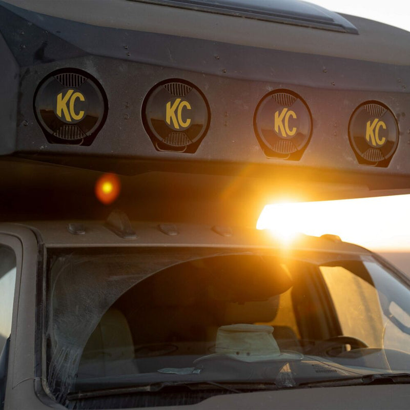 KC HiLiTES SlimLite 8" LED Light Cover & Shield on a vehicle: four round KC lights with black covers and yellow KC logos mounted above a camper truck windshield at sunset