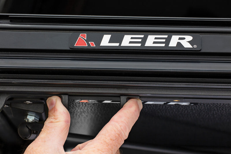Close-up of the LEER HF350M hard folding tonneau cover: latch release being pulled under a LEER-badged cover rail, close-up