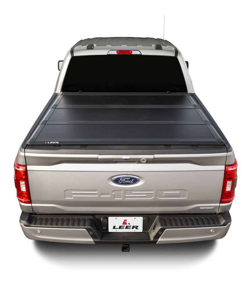 LEER HF350M hard folding tonneau cover on a pickup bed: rear view of a black hard tri-fold cover closed over a silver pickup bed, LEER plate on bumper