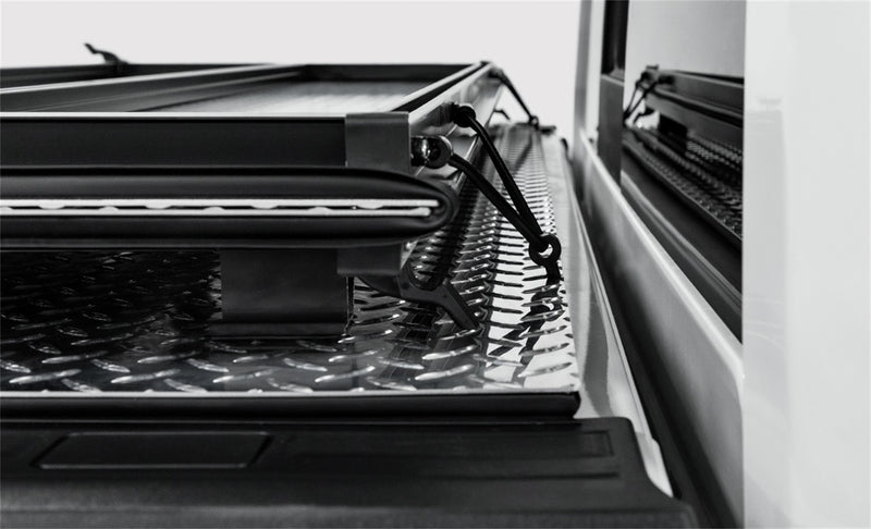 LOMAX Hard Tri-Fold tonneau cover: black-and-white close-up of a folded panel stack resting on the rear panel, secured with a bungee cord