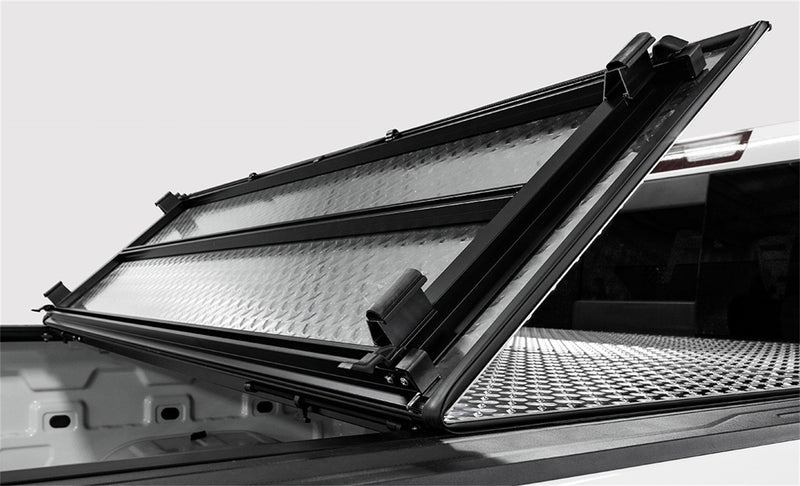 LOMAX Hard Tri-Fold tonneau cover: black-and-white photo of a white truck bed with the tri-fold panels hinged up, showing the diamond-plate aluminum underside and frame hardware