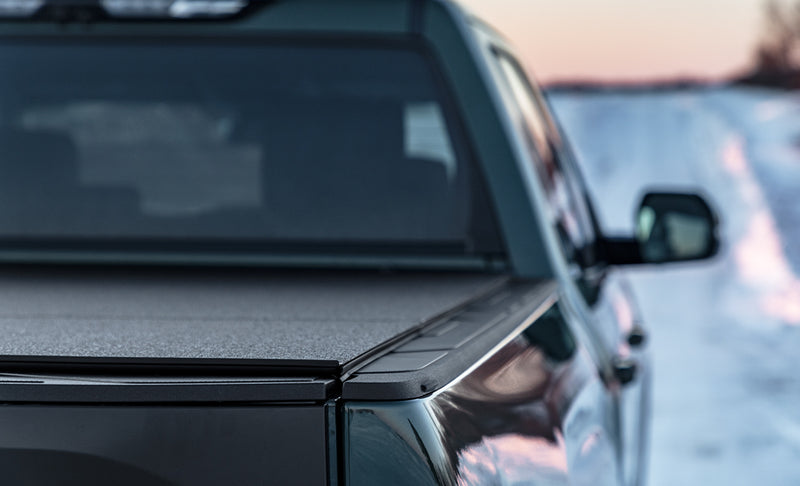 LOMAX Hard Tri-Fold tonneau cover: close rear-3/4 of a dark pickup at dusk in snow, closed textured-black tri-fold