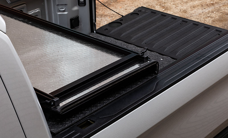 LOMAX Hard Tri-Fold tonneau cover: folded tri-fold panel with its silver diamond-plate underside up, silver truck, tailgate down with liner, carpet bed mat