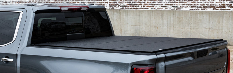 LOMAX Hard Tri-Fold tonneau cover: gray pickup side view against concrete/brick wall, tri-fold cover closed with textured black panels