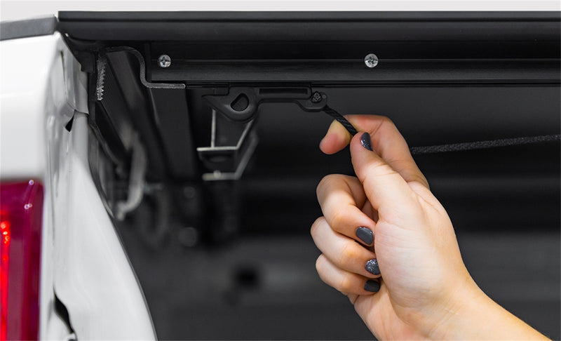 LOMAX Hard Tri-Fold tonneau cover: hand pulls cable release under the rail, white truck with red tail lamp edge
