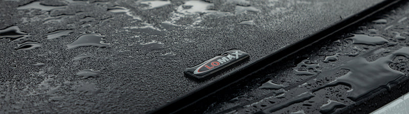 LOMAX Hard Tri-Fold tonneau cover: macro of the closed cover's textured black top with water droplets and a raised LOMAX badge near the rail