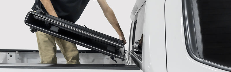 LOMAX Hard Tri-Fold tonneau cover: man lifts the folded tri-fold off a white truck, studio