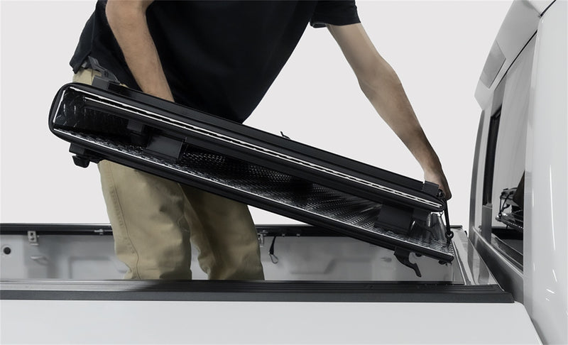 LOMAX Hard Tri-Fold tonneau cover: man lifts the folded tri-fold stack off a white truck bed, studio