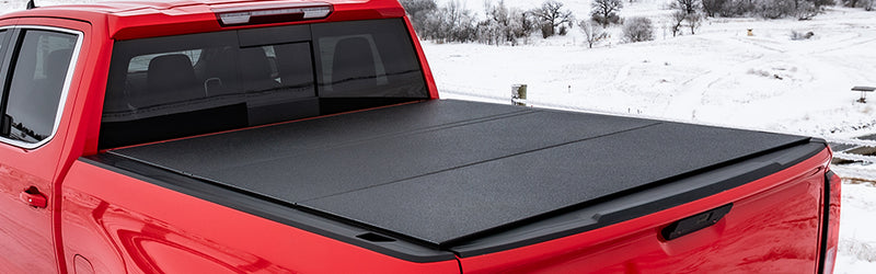LOMAX Hard Tri-Fold tonneau cover: red full-size pickup in a snowy field, tri-fold cover closed with textured black panels