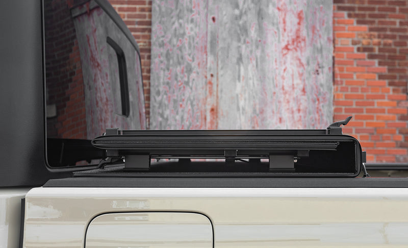 LOMAX Hard Tri-Fold tonneau cover: side view of a white bed with the tri-fold folded flat at the cab, brick wall behind