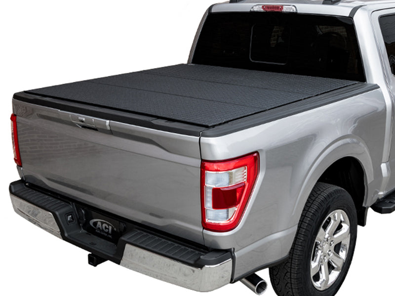 LOMAX Hard Tri-Fold tonneau cover: silver pickup on white studio background, rear 3/4 view, hard tri-fold cover closed with black diamond-pattern coated panels