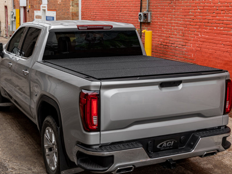 LOMAX Hard Tri-Fold tonneau cover: silver pickup parked in a brick alley, rear 3/4 view, tri-fold cover closed with black diamond-pattern coated panels