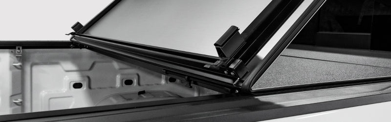 LOMAX Hard Tri-Fold tonneau cover: tri-fold panel lifted showing aluminum underside and frame clips, bare bed, monochrome studio