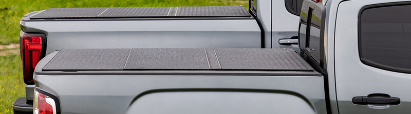 LOMAX Hard Tri-Fold tonneau cover: two gray trucks side by side on grass, both with closed tri-fold covers showing diamond-pattern coated panels