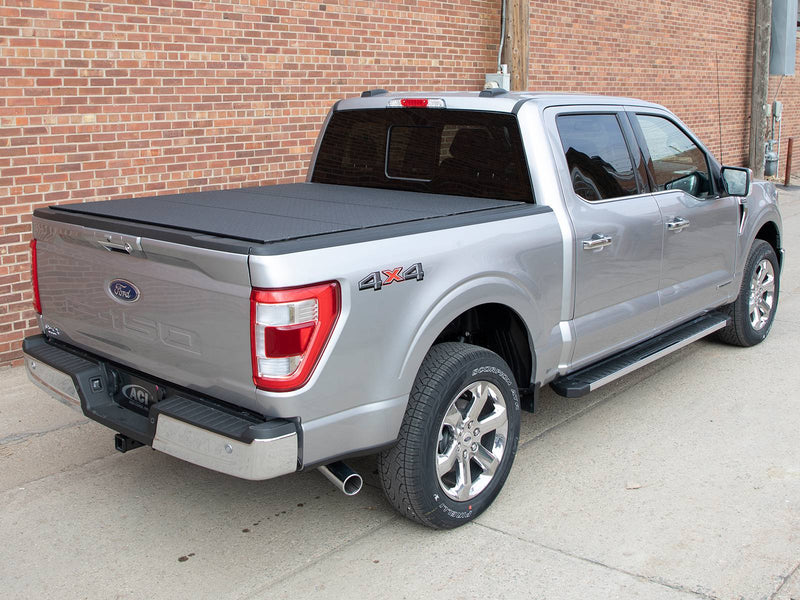 Silver pickup by a brick wall, black diamond-pattern hard tonneau cover closed, rear three-quarter view