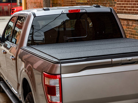 Rear three-quarter view of a pickup bed with a diamond-pattern black hard tri-fold tonneau cover closed, brick wall behind