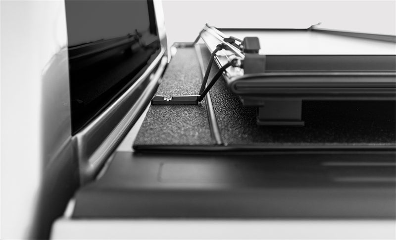 LOMAX STANCE tonneau cover: black-and-white close detail of folded panels resting on the bed rail at the cab, textured black top and bungee latch