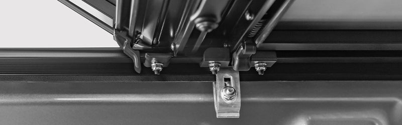 LOMAX STANCE tonneau cover: black-and-white wide close-up of the rail clamp bolted under the bed rail with folded panel hinges above