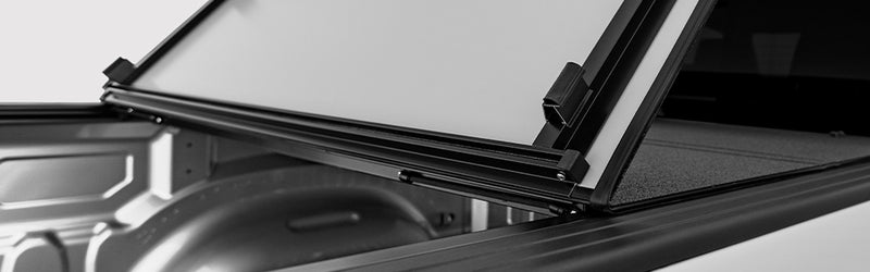 LOMAX STANCE tonneau cover: black-and-white wide detail of a panel being folded up over the bed rail, textured top visible at right