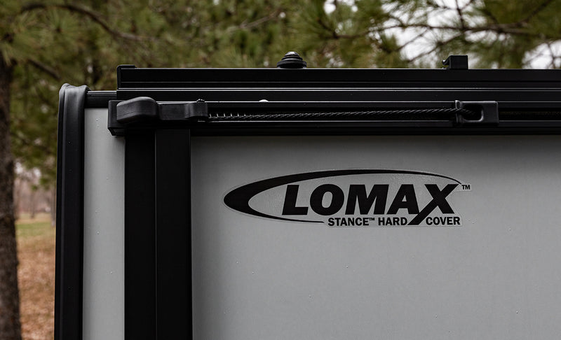 LOMAX STANCE tonneau cover: close detail of the vertical STANCE panel underside with 'LOMAX STANCE HARD COVER' decal and bungee retainer, pine background