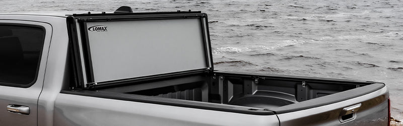 LOMAX STANCE tonneau cover: silver pickup beside water, STANCE panels vertical at cab showing gray underside with LOMAX decal, empty bed