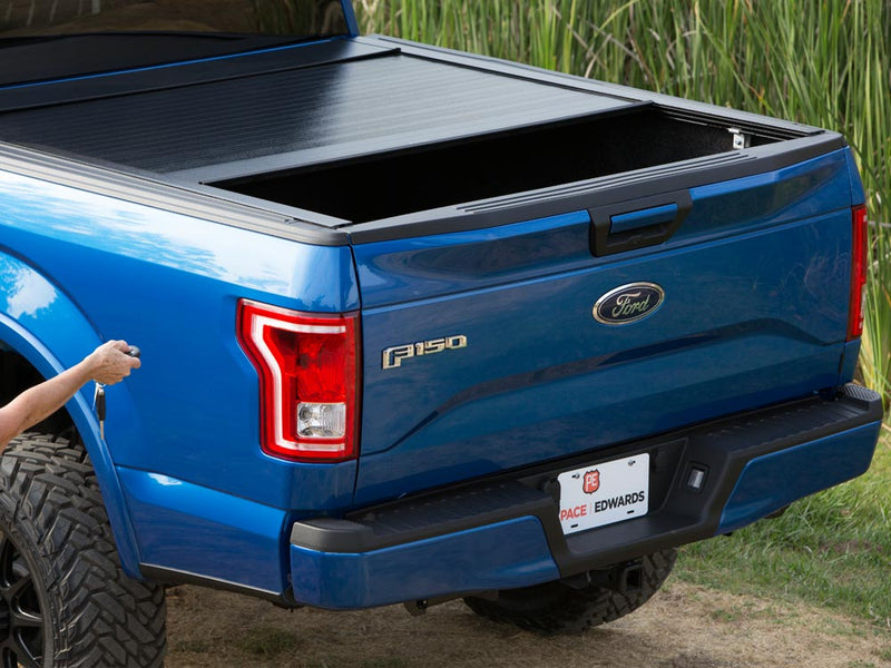 Pace Edwards BedLocker: black retractable cover partly open on a blue pickup while a hand presses the key fob