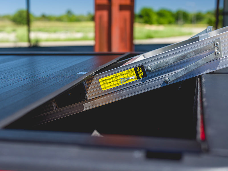 Pace Edwards BedLocker: black slatted cover with its aluminum frame tilted up, yellow caution label visible, outdoors