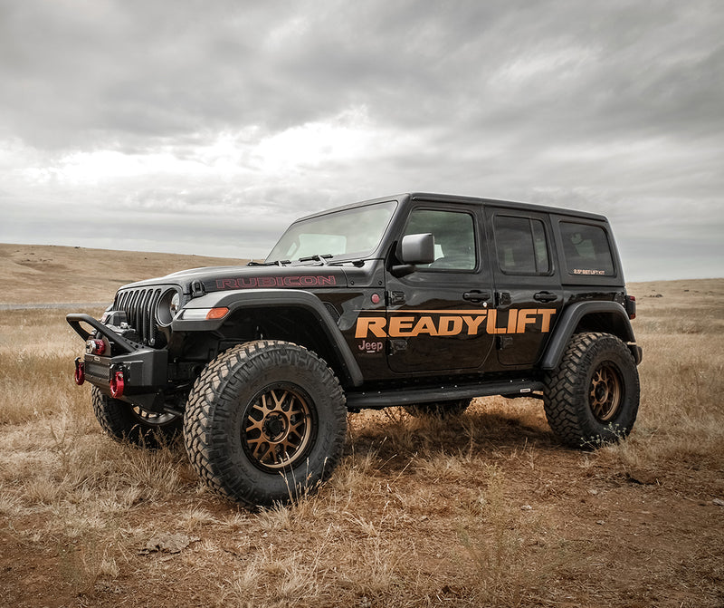 ReadyLIFT Coil Spring Lift Kit installed on a Jeep Wrangler JL: A black lifted Jeep Wrangler Rubicon four-door with ReadyLIFT graphics parked in a dry grass field