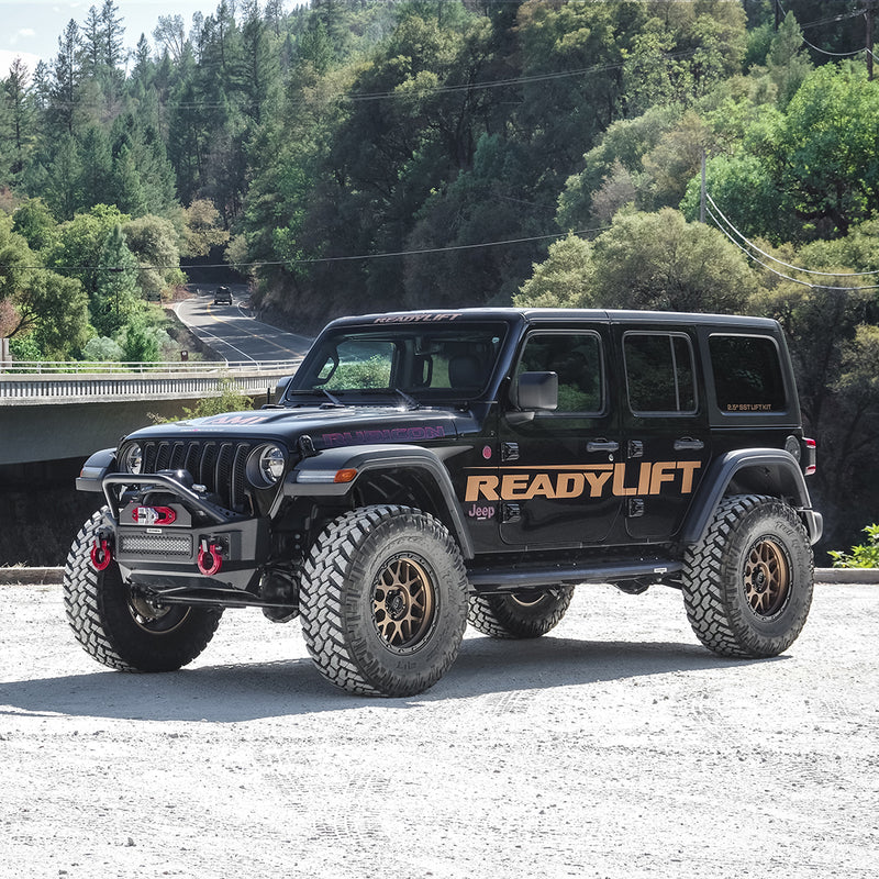 ReadyLIFT Coil Spring Lift Kit installed on a Jeep Wrangler JL: A black lifted Jeep Wrangler Rubicon four-door with ReadyLIFT graphics parked on gravel by a bridge