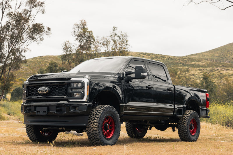 ReadyLIFT Coil Spring Lift Kit installed on a Ford F-250 Super Duty: black Ford Super Duty crew cab lifted on red wheels and mud tires in a dry hillside setting